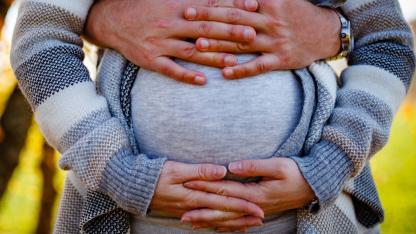 Eine schwangere Person umfasst den Bauch mit ihren Händen, von hinten wir sie von einer anderen Person umfasst.